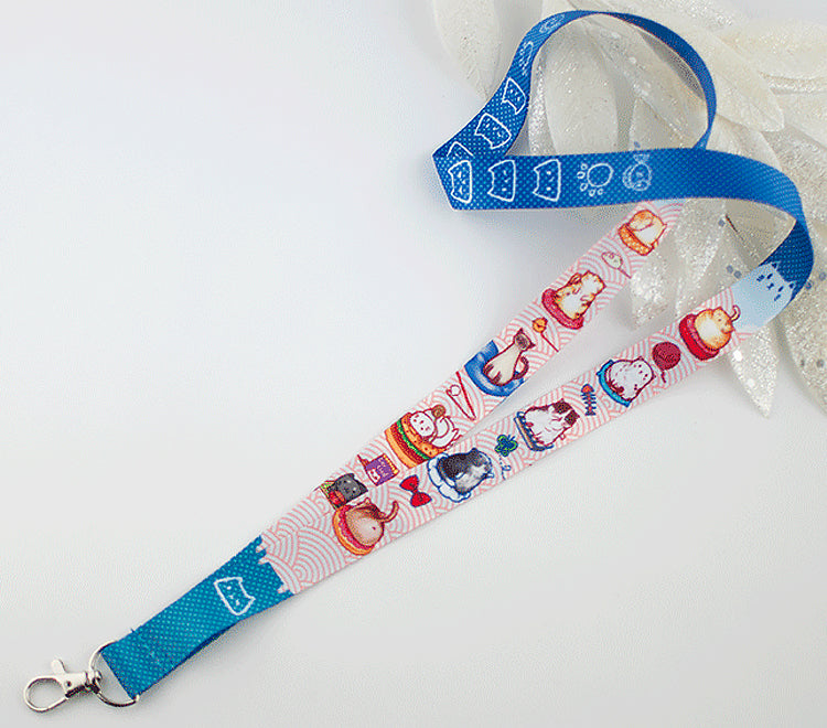 Cat lanyard with half blue dotted pattern and half light blue Japanese wave pattern. There are designs of various breeds of cats sitting and relaxing on cat cushions. For example, one tuxedo kitty is standing up and trying to catch a butterfly. A Ragdoll kitten grooming itself. A Siamese kitten being an absolute queen and sitting gracefully. A gray kitten enjoying a bowl of cat snack from its human butler. The lanyard have a lobster claw clasp to hold your badges.